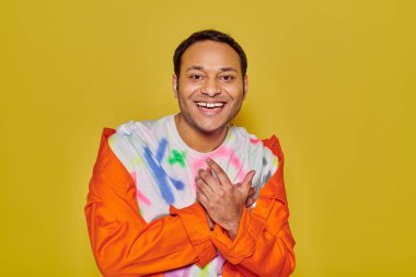 joyous indian man in orange jacket and diy t-shirt smiling and looking at camera on yellow backdrop clipart