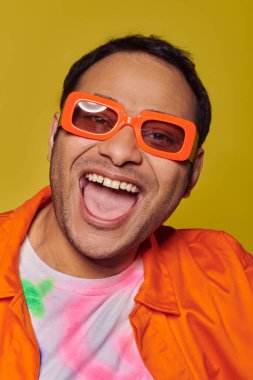 self-expression concept, excited indian man in orange sunglasses smiling on yellow backdrop clipart