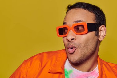 self-expression concept, surprised indian man in orange sunglasses looking away on yellow backdrop clipart