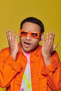 confused indian man in orange sunglasses looking away and gesturing on yellow background, expressive clipart