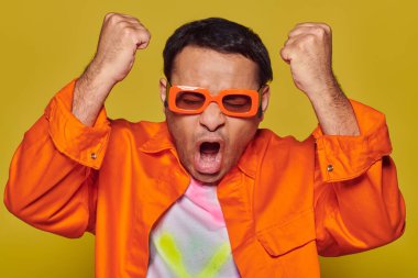 emotional winner celebrating victory, indian man in orange sunglasses gesturing on yellow background clipart