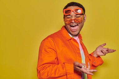 funny face, excited indian man trying on different trendy sunglasses and smiling on yellow backdrop clipart