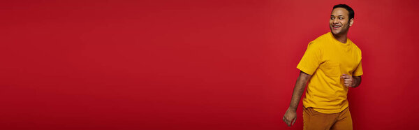 gleeful indian man in bright casual clothes looking away and smiling on red background, banner