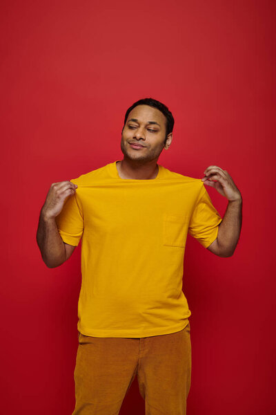 Модный индийский мужчина трогает яркую футболку и смотрит в сторону на красный фон, личный стиль