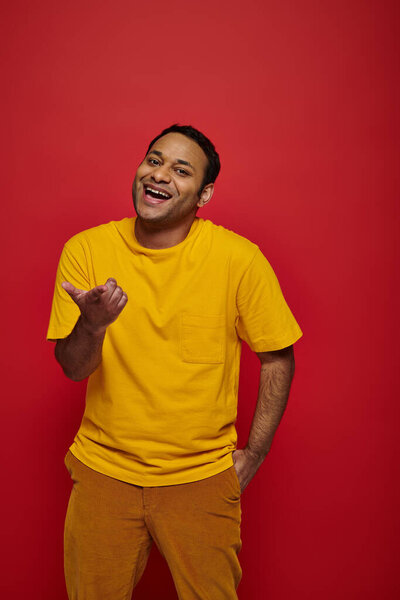cheerful indian man in bright casual clothes mocking and pointing with finger on red background