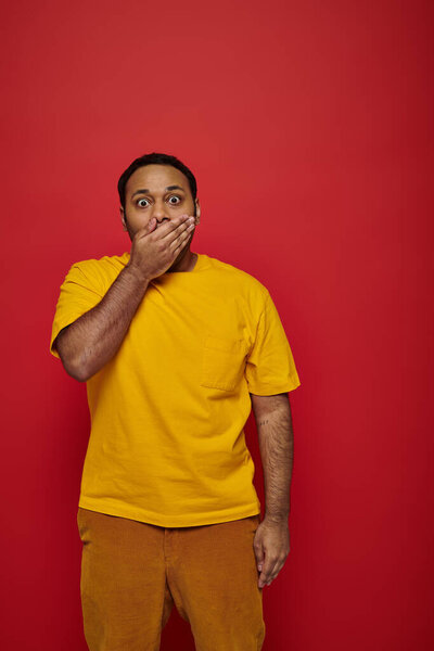 shocked indian man in bright clothes covering mouth and looking at camera on red background