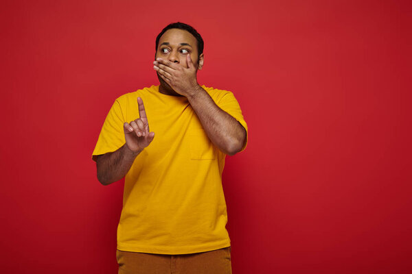 shocked indian man in bright clothes covering mouth and showing finger on red background, warning