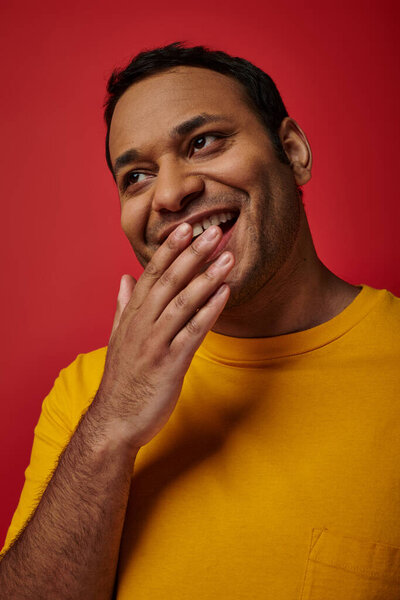 shy indian man in yellow t-shirt smiling and covering mouth with hand on red background in studio