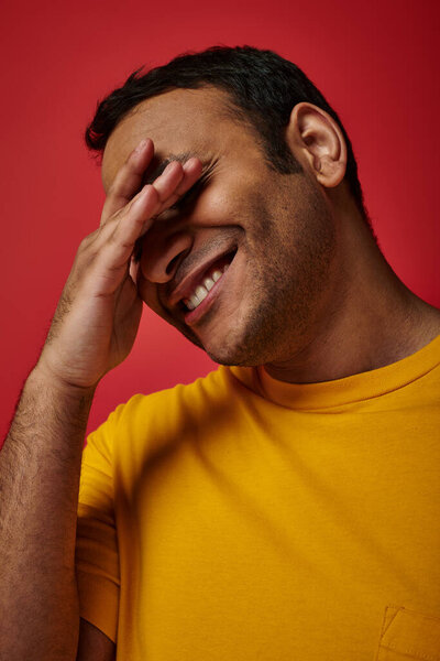 shy indian man in yellow t-shirt smiling and covering eyes with hand on red background in studio