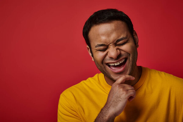 joyous indian man in yellow t-shirt smiling with closed eyes on red backdrop in studio, portrait