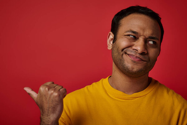 skeptical indian man in yellow t-shirt pointing with thumb and looking away on red backdrop