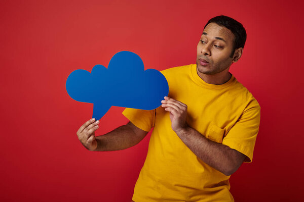 indian man in yellow t-shirt looking at blank thought bubble on red backdrop, upset face