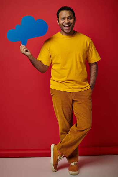 happy man in yellow t-shirt holding blank thought bubble on red backdrop, hand in pocket pose