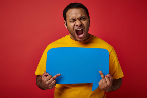 provocative indian man holding blank speech bubble and showing middle fingers on red background