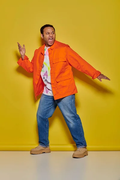 オレンジのジャケットとジーンズの感情的なインドの男は 黄色い背景にジェスチャーと叫びます — ストック写真