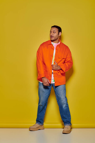 full length of stylish indian man in orange jacket and denim jeans standing on yellow backdrop