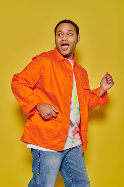 portrait of amazed indian man in orange jacket running away and looking back on yellow backdrop