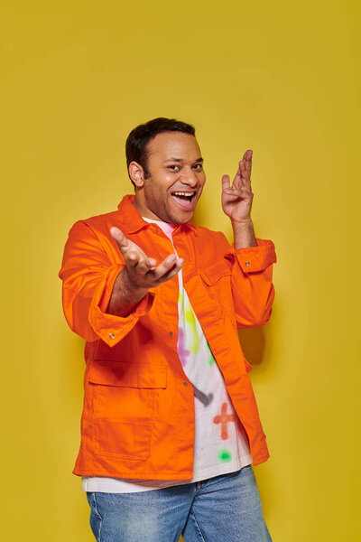 portrait of excited indian man in orange jacket and diy t-shirt gesturing on yellow backdrop