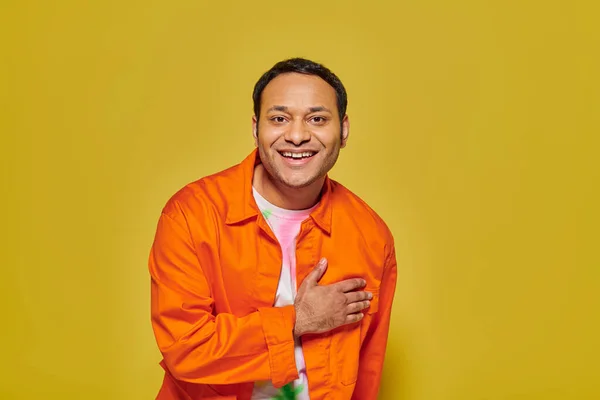 stock image portrait of joyful indian man in orange jacket looking at camera and smiling on yellow backdrop