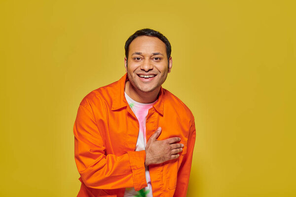 portrait of joyful indian man in orange jacket looking at camera and smiling on yellow backdrop
