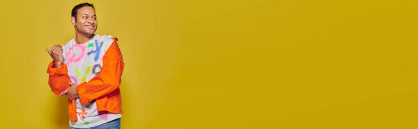 positive indian man in orange jacket and diy t-shirt pointing with thumb on yellow backdrop, banner