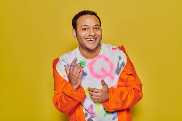 gleeful indian man in orange jacket and diy t-shirt smiling and looking at camera on yellow backdrop