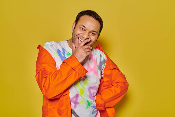 cheerful indian man in orange jacket and diy t-shirt smiling on yellow backdrop, cover mouth