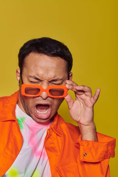 face expression, indian man adjusting orange sunglasses and grimacing on yellow backdrop, attitude
