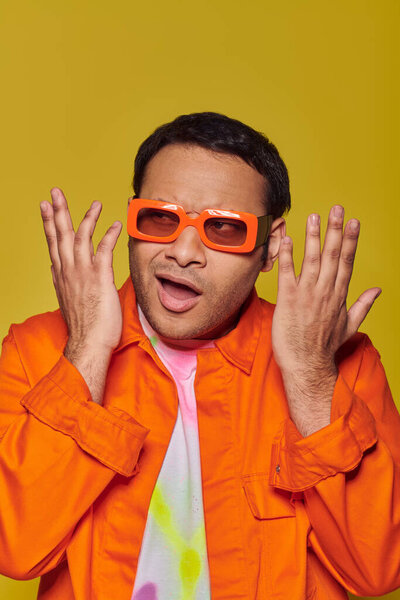 confused indian man in orange sunglasses looking away and gesturing on yellow background, expressive