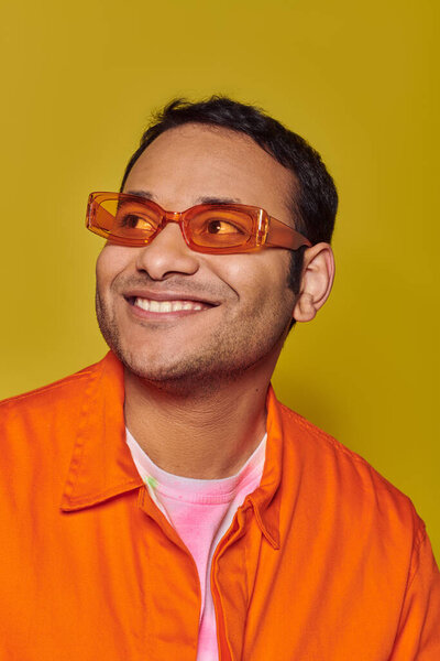 happy indian man in orange sunglasses looking away while smiling on yellow background, side glance