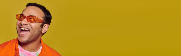 positive indian man in orange sunglasses looking away and smiling on yellow background, banner