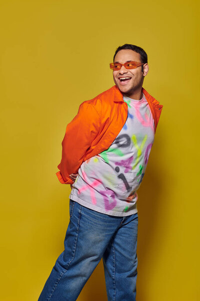 amazed indian man in sunglasses and vibrant attire looking away and smiling on yellow background