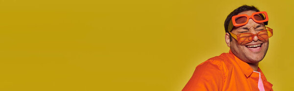 stylish indian man trying on different trendy sunglasses and smiling on yellow backdrop, banner