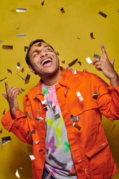 excited indian man in orange jacket gesturing near confetti on yellow backdrop, party concept