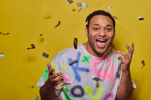 счастливый индиец в модной футболке жестикулируя на фоне падающих конфетти на желтом фоне, концепция вечеринки