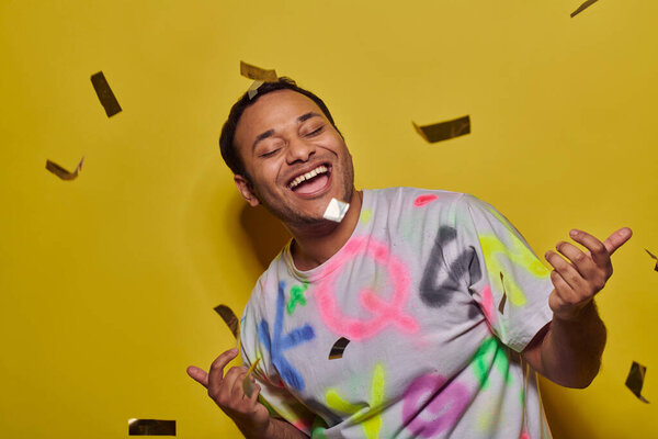 возбужденный индийский мужчина в футболке жестикулируя на фоне падающих конфетти на желтом фоне, концепция вечеринки