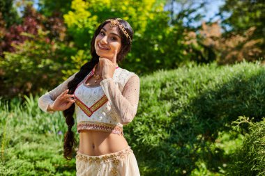 Zarif etnik giyimli neşeli Hintli kadın yeşil yaz parkında kameraya poz veriyor.
