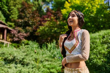 Yaz parkında yeşilliğin yanında gülen geleneksel kıyafetli heyecanlı Hintli kadın.