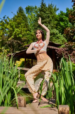 carefree and graceful indian woman in ethnic attire dancing in summer park on sunny day clipart