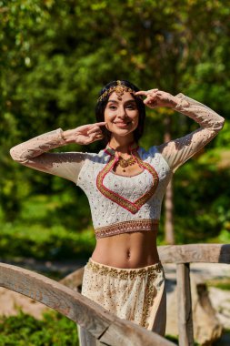 Parkta güneşli bir günde geleneksel kıyafetli şık Hintli kadın kameraya poz veriyor ve gülümsüyor.