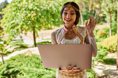 Yaz parkında bilgisayar başında el sallayan geleneksel giyinmiş neşeli Hintli kadın.