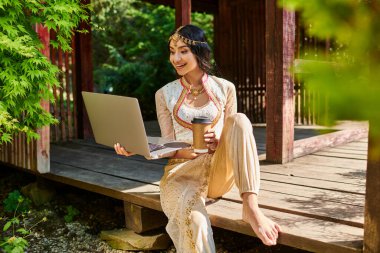Etnik giyimli mutlu Hintli kadın dizüstü bilgisayarlı ve parktaki ahşap bölmede kahve alıyor.