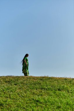summer, ethnic heritage, young indian woman under blue cloudless sky in green field clipart