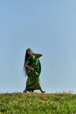 Yaz tatili, kaygısız Hintli kadın sari ile yeşil çayırlarda yürüyor mavi bulutsuz gökyüzünün altında