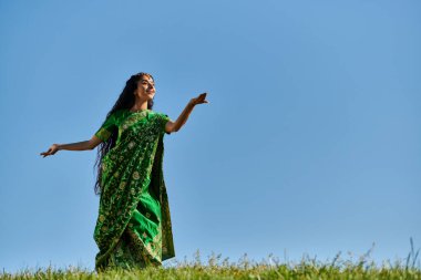 Geleneksel sari giymiş zarif Hintli kadın mavi gökyüzünün altında yeşil çayırlarda dans ediyor.