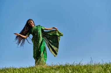 Mavi gökyüzünün altındaki yeşil tarlada geleneksel giyinmiş neşeli Hintli kadının yaz dansı.