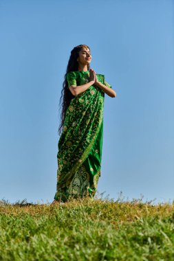 Mutlu Hintli kadın sari içinde dua eden elleri ve mavi gökyüzünün altındaki çimlerde kapalı gözleri olan, yaz günü