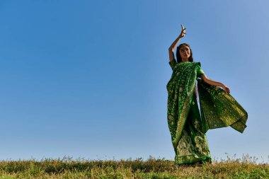 Mavi gökyüzünün altındaki yeşil alanda otantik sari giymiş gülümseyen Hintli kadının yaz dansı.