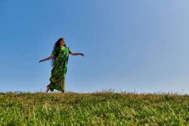 Canlı bir yaz, etnik giysiler içinde neşeli bir Hintli kadın mavi gökyüzünün altında yeşil alanda koşuyor.