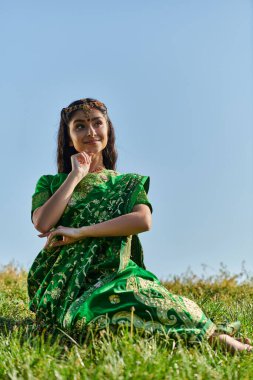 Arka planda gökyüzü olan çimenli tepede poz veren geleneksel sari giymiş pozitif Hintli kadın.